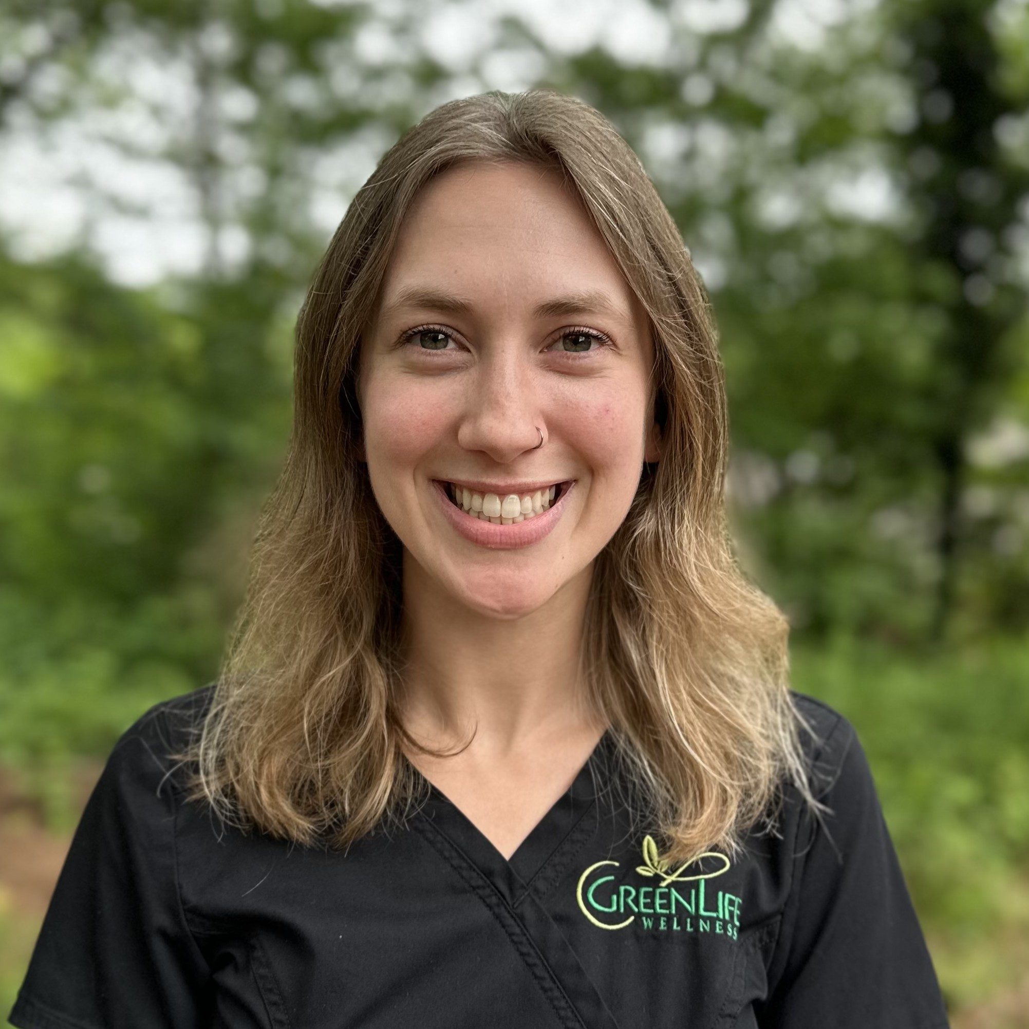 Chiropractic Greenville SC Leah Coffey Meet The Team Leah Coffey smiling for photo outside with trees in background and wearing black scrubs